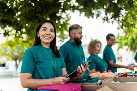 A smiling woman with a tablet is volunteering at a community event with a group of people sorting supplies in the background. | Medi-Weightloss