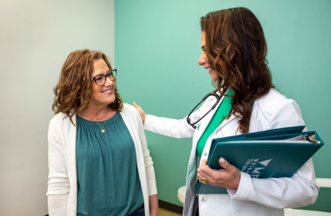 A doctor places a reassuring hand on the shoulder of a smiling middle-aged woman with curly hair and glasses. The doctor holds a teal Medi-Weightloss folder. | Medi-Weightloss