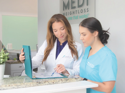 A female doctor in a white coat points to a page in a binder as a female nurse in blue scrubs observes. | Medi-Weightloss